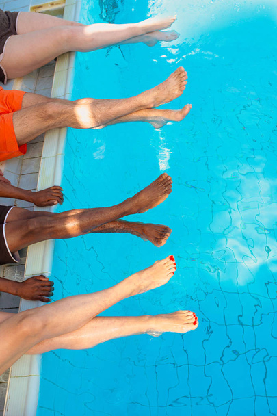 ноги четырех друзей (простых людей) разных рас, сидящих у бассейна в солнечный день. Вечеринка Саммер. Концепция отдыха и спорта
. - Фото, изображение