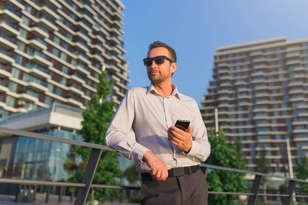 Exitoso ejecutivo de negocios utilizando el teléfono inteligente al aire libre. Edificios de oficinas en segundo plano
. - Foto, Imagen