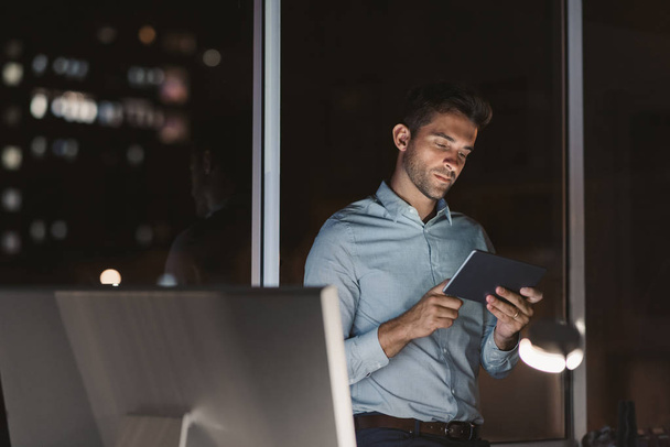 fokussierter junger Geschäftsmann, der spät in der Nacht allein in einem Büro steht und mit einem digitalen Tablet mit Stadtbeleuchtung im Hintergrund arbeitet - Foto, Bild