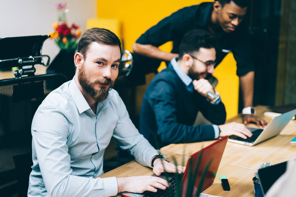 Group of young it professionals developing program code for antivirus on laptop computer enjoying collaboration indoors, portrait of successful man looking at camera while colleagues talking near - Foto, Imagen
