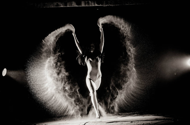 das Mädchen mit dem Mehl auf dem Körper streckt die Arme mit geworfen Mehl auf schwarzem Hintergrund Schwarz-Weiß-Bild - Foto, Bild