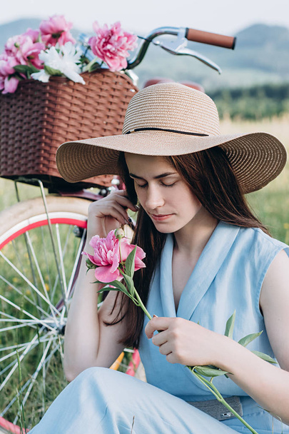 花のバスケットで自転車の近くにピンクのピオニーを持つ帽子をかぶった美しい女性のクローズアップ肖像画。公園で白人女性のリラクゼーション。コピー、空き領域 - 写真・画像