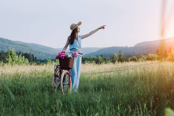 Outdoor portrait of young happy smiling woman in hat and enjoys mountain nature during the sunset. She holds a bicycle with a basket of flowers. Concept outdoor nature lifestyle - Photo, image