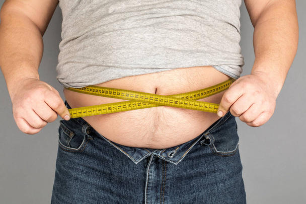 A man measures his fat belly with a measuring tape. on a gray ba - Photo, Image