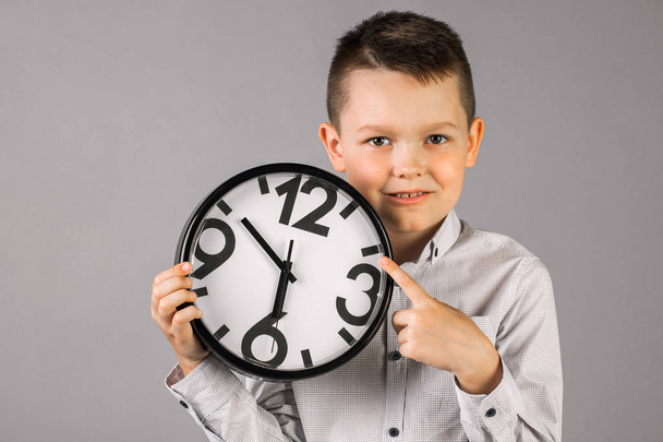 αγόρι φορώντας ένα καρό πουκάμισο και κρατώντας ένα ρολόι, σε ένα γκρίζο τοίχο BA - Φωτογραφία, εικόνα