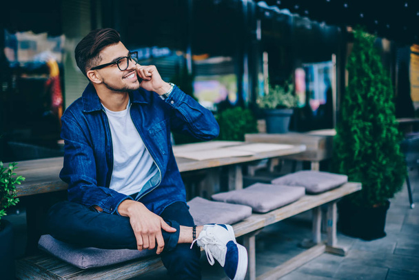 Cheerful caucasian male in spectacles having mobile phone conversation sitting outdoors, happy hipster guy in trendy wear satisfied with connection in roaming  making telephone call and smiling - Photo, Image