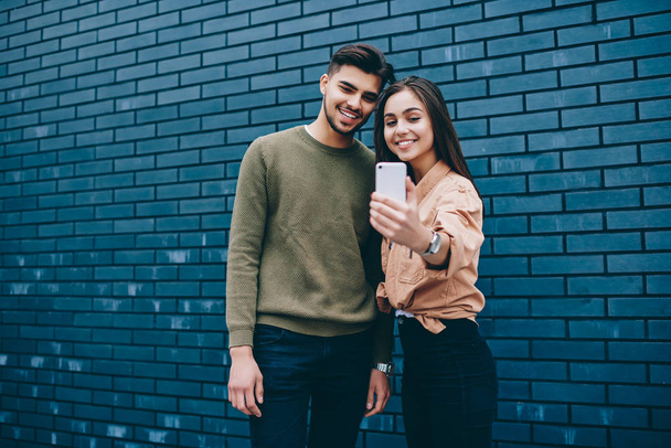Cheerful couple in love  dressed in casual posing for selfie on smartphone camera standing together,young smiling teen hipster girl and guy using modern mobile phone application making common picture - Foto, Bild