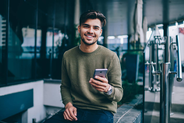 Portrait of cheerful handsome male using mobile phone and wireless internet for chatting and networking , young hipster guy looking at camera satisfied with good 4G connection for sending messages - Φωτογραφία, εικόνα