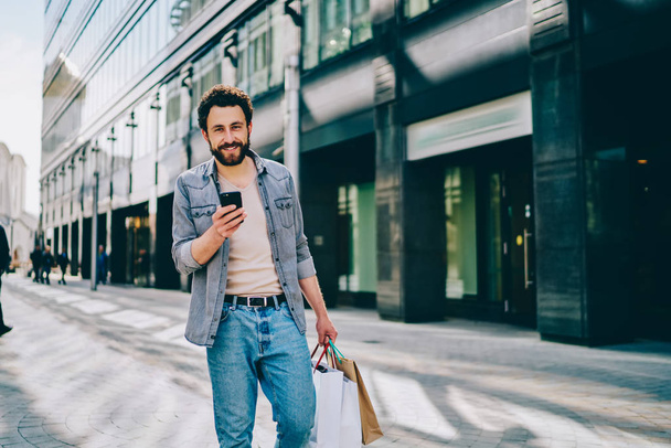 Portré boldog csípő srác gazdaság okostelefon a kezében, miközben mosolyogva a kamera és a gyaloglás közelében bevásárlóközpont csinál vásárolni fekete péntek. Vidám kaukázusi vendég papírzsákokban a boltokban - Fotó, kép