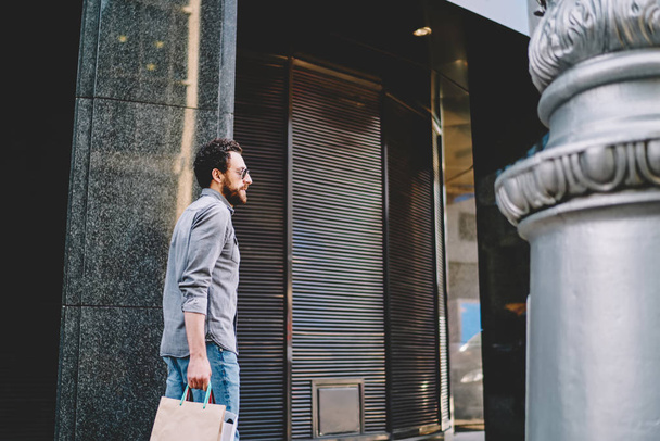 Young man customer in stylish sunglasses doing shopping in black friday with big dales in modern mall.Stylish hipster guy with paper bags with fashionable goods strolling in urban setting - 写真・画像