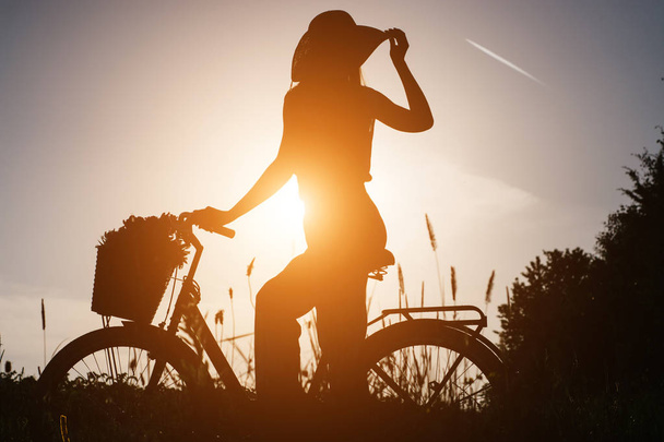 Image of silhouette of woman outdoor sitting on vintage bicycle with a basket of flowers and enjoying sunset. Active vacation concept. - Photo, Image