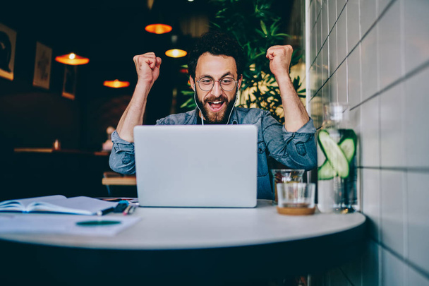 Overjoyed hipster guy in earphones cheers for team watching match on laptop computer during free time in cafe interior, amazed emotional male freelancer celebrating achievement and completing projec - Фото, зображення