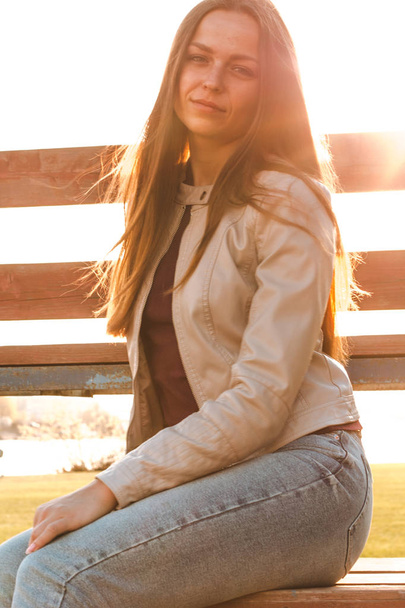 uzun saçlı güzel kız bej deri ceket ve kot güneş ışığı karşısında oturur - Fotoğraf, Görsel