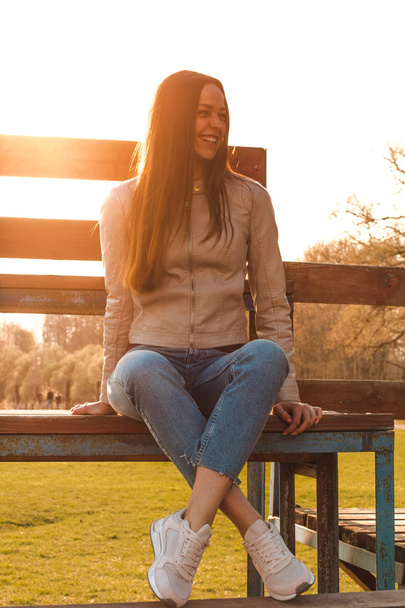 mooi meisje met lang haar zit tegenover het zonlicht in een beige leren jas, jeans en sneakers lachen veel - Foto, afbeelding
