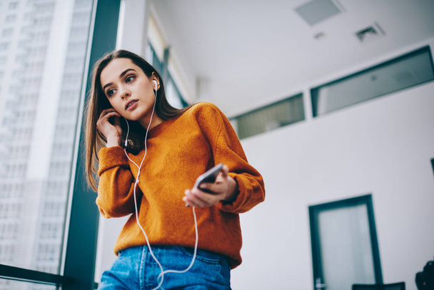 Pondering caucasian hipster girl dressed in casual look using wireless 4g connection for listening radio online via smartphone application, thoughtful woman in electronic headphones enjoying song - Photo, Image