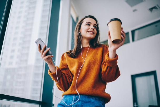 Pozytywna Hipster dziewczyna uśmiechnięta podczas słuchania książki audio motywacji przez elektroniczne słuchawki i aplikacji online dla edukacji trzymając kawę, aby przejść w pomieszczeniu, szczęśliwa kobieta korzystających czas na muzykę - Zdjęcie, obraz
