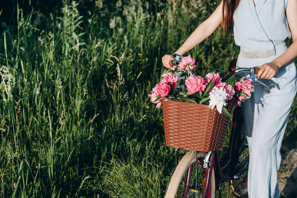 Close up shot of young beautiful girl, she holds a bicycle with a basket of red and white peonies flowers. Bike with basket full of red and white flowers. Active vacation concept. - Фото, зображення