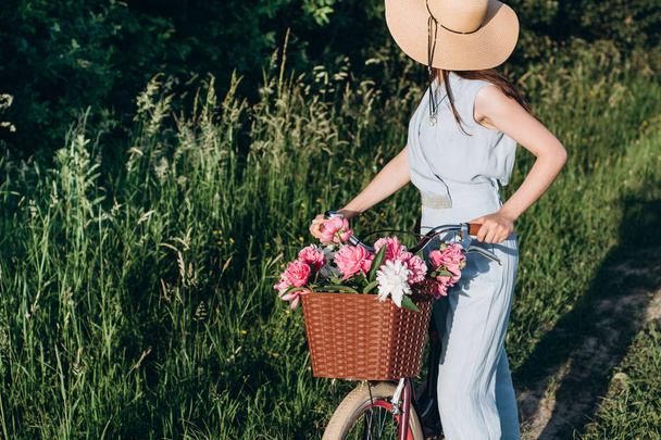 Horizontal portrait of young beautiful girl, she holds a bicycle with a basket of red and white peonies flowers. Bike with basket full of red and white flowers. Active vacation concept. - Photo, Image