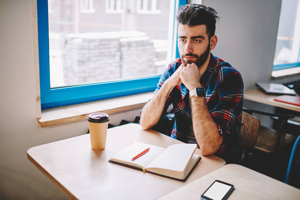 Serious bearded hipster student looking straight while sitting at desktop with notebook and thinking on homework in academy.Young man in casual wear studying and preparing for seminar in college - Foto, Imagem