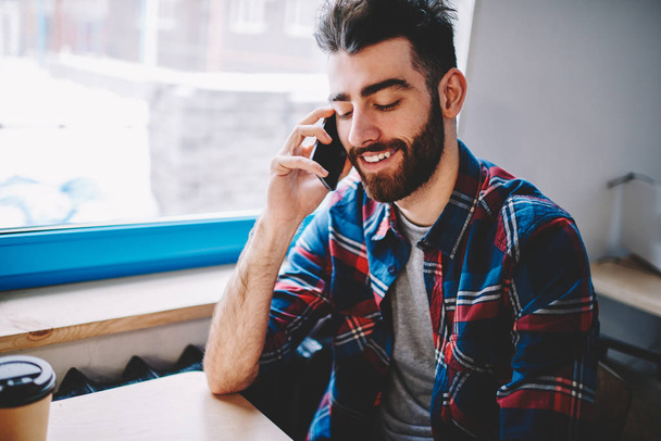 Cheerful bearded young man talking with friend on smartphone using high speed mobile connection.Positive student calling on mobile phone and communicating with operator sitting indoors - Foto, Imagem