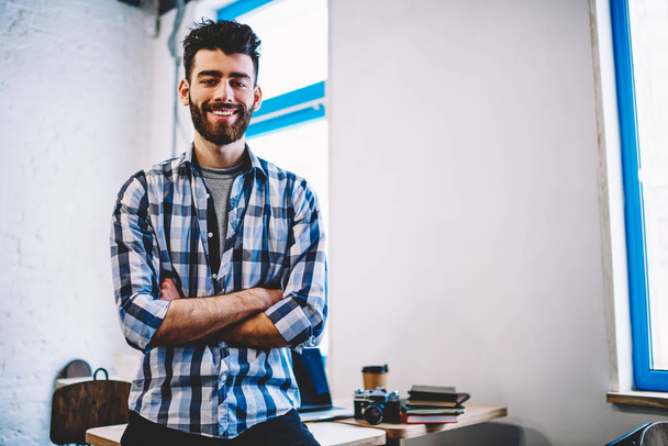 Half length portrait of successful bearded young man in casual wear smiling at camera.Positive hipster guy with crossed hands standing in own modern design studio office interior feeling confident  - Fotografie, Obrázek
