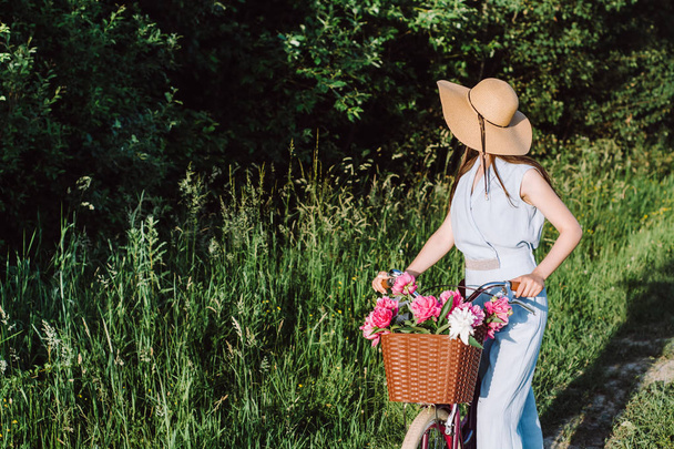 Beautiful and young girl in a hat rides a bicycle with a basket of red and white flowers on the background of nature. Concept outdoor nature lifestyle. - Fotografie, Obrázek