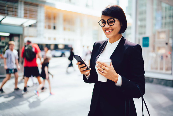 Half length portrait of cheerful successful female owner of company in optical spectacles smiling and looking at camera while holding modern cellular phone and takeaway coffee in hands during break - Фото, изображение