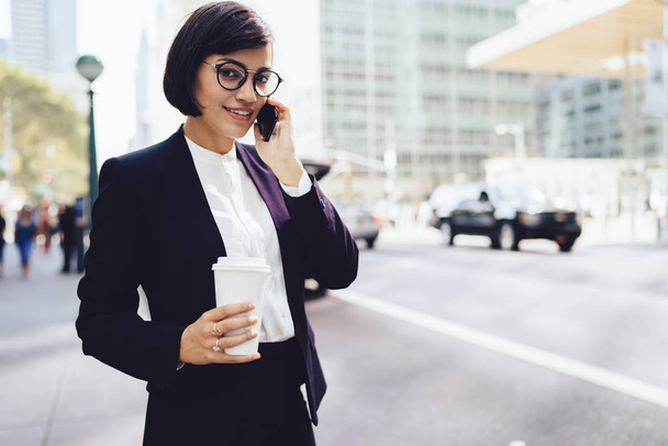 Successful female entrepreneur in eyewear discussing financial news on mobile phone during work break with coffee in hands, smiling businesswoman talking with partner via application on smartphone - Zdjęcie, obraz