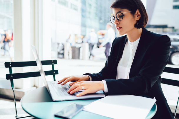 Concentrated female lawyer in optical eyeglasses booking tickets on website while sitting with modern laptop device with wireless internet connection, concept of business people and technology - Photo, image