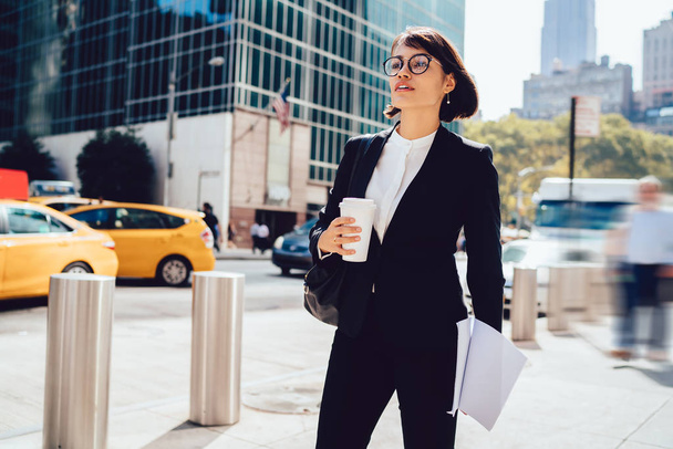 Уверенная деловая женщина в формальной одежде спешит в офис компании на Манхэттен-стрит, брюнетка-менеджер в стильных очках с кофе на руках и бумажными документами для проекта
 - Фото, изображение