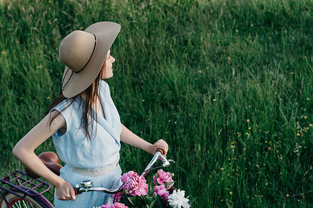 Outdoor portrait of joyful girl in straw hat sitting on bicycle on nature background. Bike with basket full of red and white flowers. Active vacation concept. - Foto, immagini