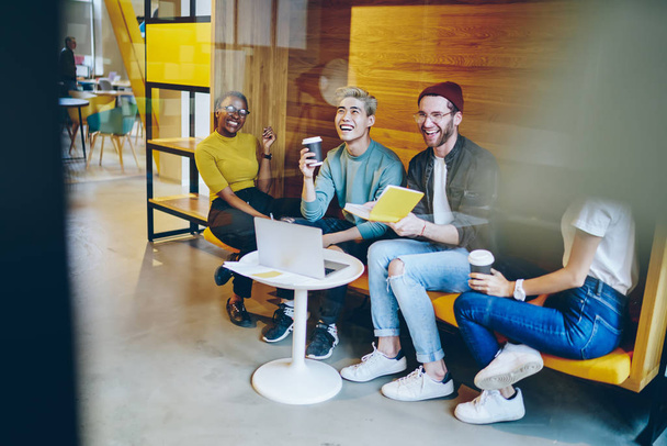 Cheerful multicultural young people laughing during coffee break in modern office.Positive diverse hipster students with books in hands communicating with each other and having fun - 写真・画像
