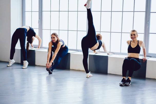 Positive attractive girls in trendy sportswear communicated and doing stretch exercises before training in dancing class, group of young active fitness female friends enjoying time at sport club - Foto, Imagem