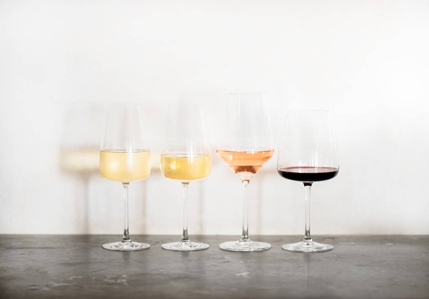 Variety of wine types. Light and full-bodied white, rose and red wine in glasses in row over concrete table, white wall background, copy space. Wine list for cafe, wine boutique or degustation - Photo, image