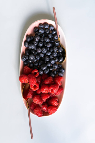Summer berries blueberries, raspberries on the new elegant plates of pink color with teaspoons. New luxury Golden cutlery view from above on a isolated white background. Top view. - Foto, Bild