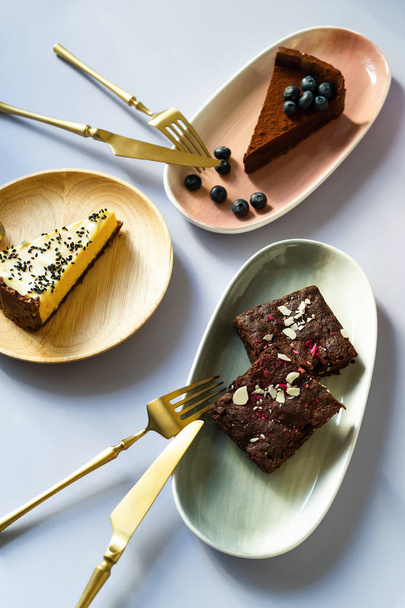Slices of cake desserts with berries and nuts on plates with different colors, pink, green, wooden material. Golden Fork with a knife for eating. Chocolates, brownies, cheesecake. - Foto, Imagem