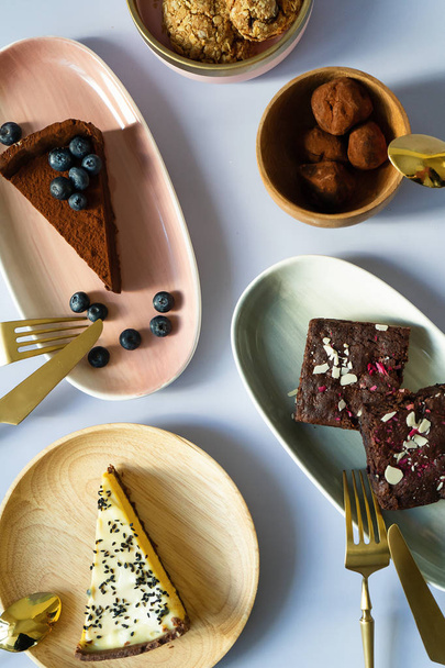 Slices of cake desserts with berries and nuts on plates with different colors, pink, green, wooden material. Golden Fork with a knife for eating. Tea spoon. Chocolates, brownies, cheesecake, biscuits. - Foto, Bild