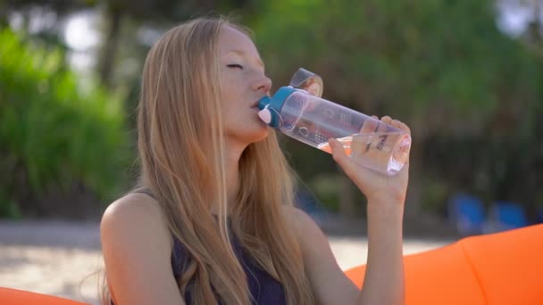 Superslowmotion shot of a young woman on a tropical beach sits on an inflatable sofa and drinks water from a multi useable plastic bottle. Drink more water. Reusable plastic and Reduce plastic waste - Filmati, video