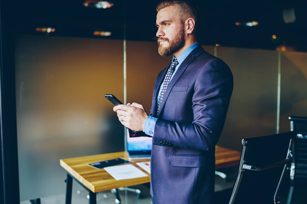 Confident entrepreneur dressed in suit chatting online on modern smartphone device while standing in office inteior, Serious businessman messaging on cellular, checking online banking via application - Photo, Image