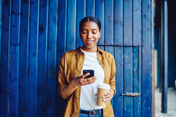 Jovem afro-americana alegre segurando café para ir e conversar on-line no telefone celular usando internet 4G pública de pé contra a parede em ambiente urbano.Blogueiro hipster de pele escura positiva - Foto, Imagem