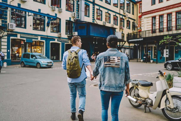 Back view of dark skinned girlfriend and caucasian boyfriend tourists walking together on street of beautiful city.Diverse couple in love holding hands each other and traveling in new modern town - Fotoğraf, Görsel