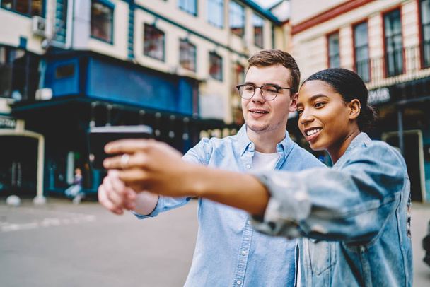 Happy multicultural young marriage making selfie picture on front camera of smartphone standing in urban setting.Positive dark skinned hipster girl with caucasian friend making photo on phone for blog - Photo, Image