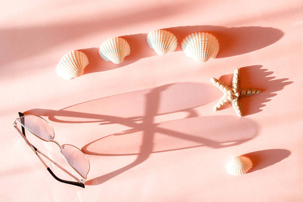 Женские розовые очки с металлической рамой отбрасывают длинную тень на розовую поверхность с морской звездой и раковиной
.  - Фото, изображение