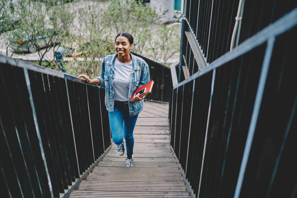 Portrait of cheerful african american young woman dressed in denim casual wear holding textbook with smartphone and climb the stairs.Positive beautiful dark skinned hipster girl smiling at camera - Valokuva, kuva