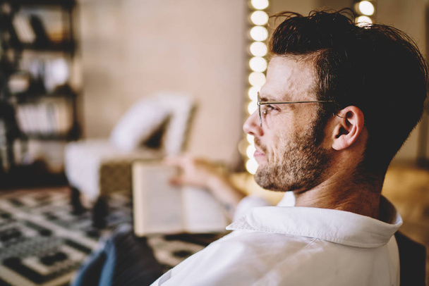 Side view of intelligent caucasian hipster guy in optical eyewear looking away and dreaming during leisure time in home apartment, handsome man thinking about youth lifestyle while taking rest indoors - Foto, immagini