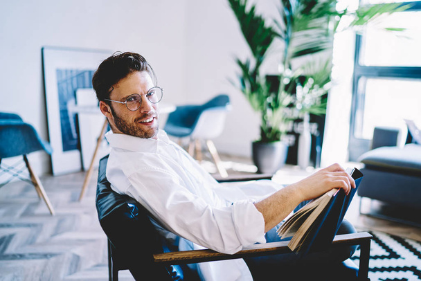 Portrait of positive hipster guy in stylish spectacles for provide eyes protection sitting in chair at home interior and feeling comfort and enjoyment from spending leisure time for literature hobby - Photo, image