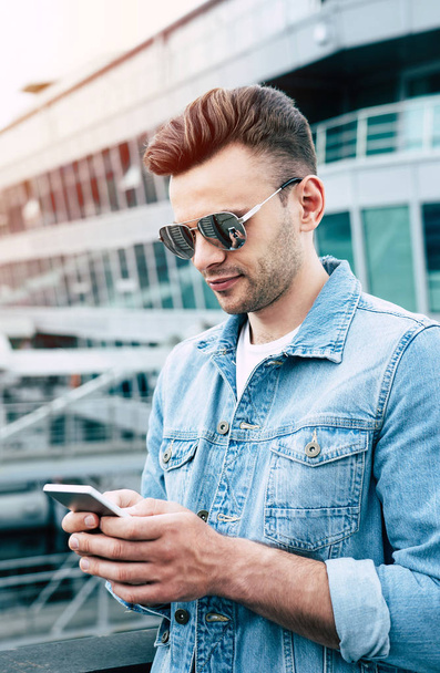 Navigare in rete. Moderno sorridente bell'uomo alla moda in occhiali da sole e jeans usura sta utilizzando un telefono all'aperto in città
. - Foto, immagini