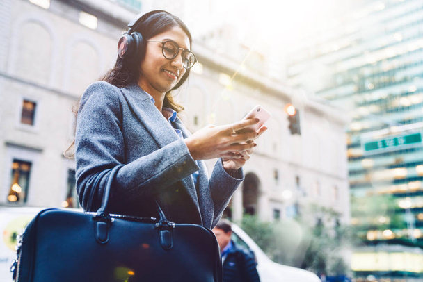 Smiling Indian female listening to music in headphones while standing on city street and relaxing after work. Young woman enjoying playlist in earphones. Watching content in social network, millennial - Φωτογραφία, εικόνα