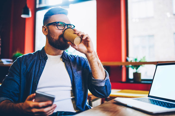Caucasian hipster guy drinking caffeine beverage from paper takeaway cup spending time in coffee shop for communicate via smartphone and laptop computer with copy space area for advertising text - Photo, image