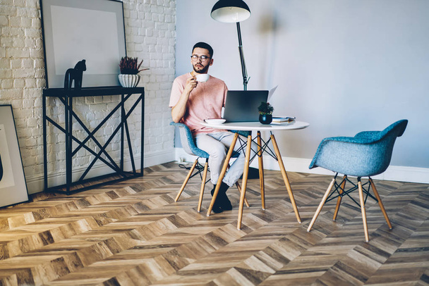 Pondering hipster guy enjoying coffee break during working remotely on laptop device in home interior with stylish design, contemplative male copywriter thinking about idea for new article on web page - Fotografie, Obrázek